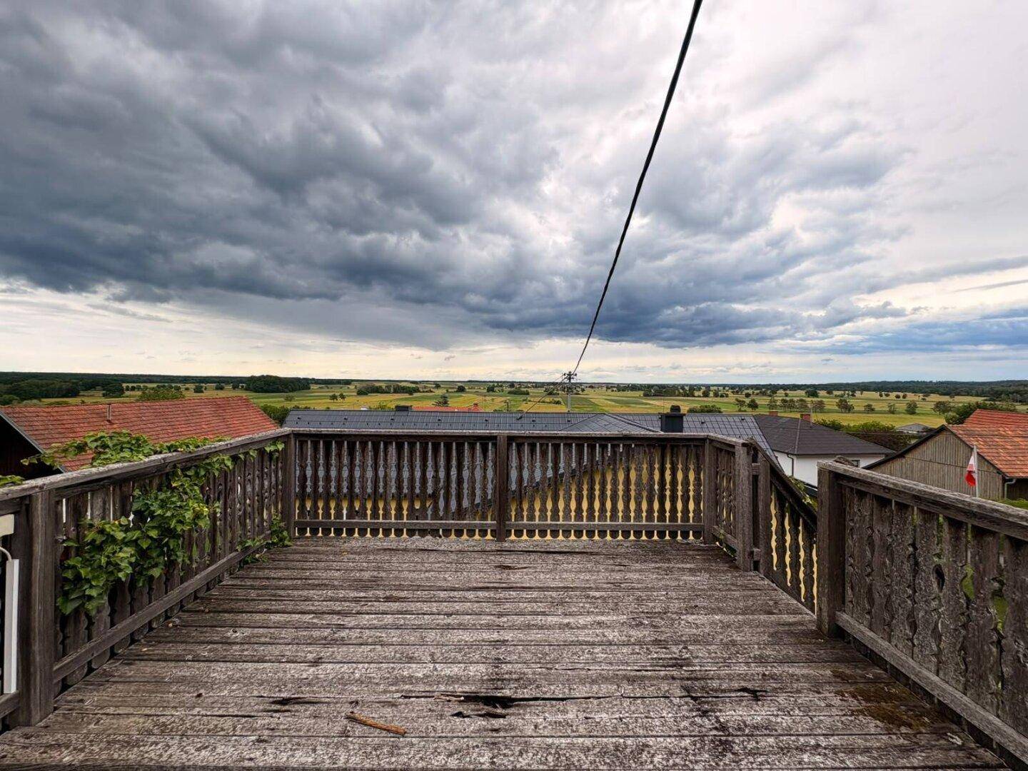 Weitblick von einer Holzterrasse über eine ausgedehnte grüne Landschaft unter dramatischem Himmel.