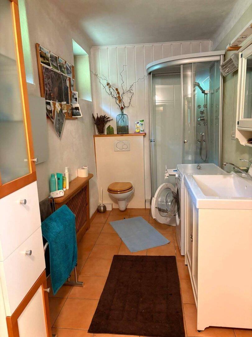 Badezimmer mit Dusche, WC, Waschmaschine und Terrakotta-Fliesenboden.