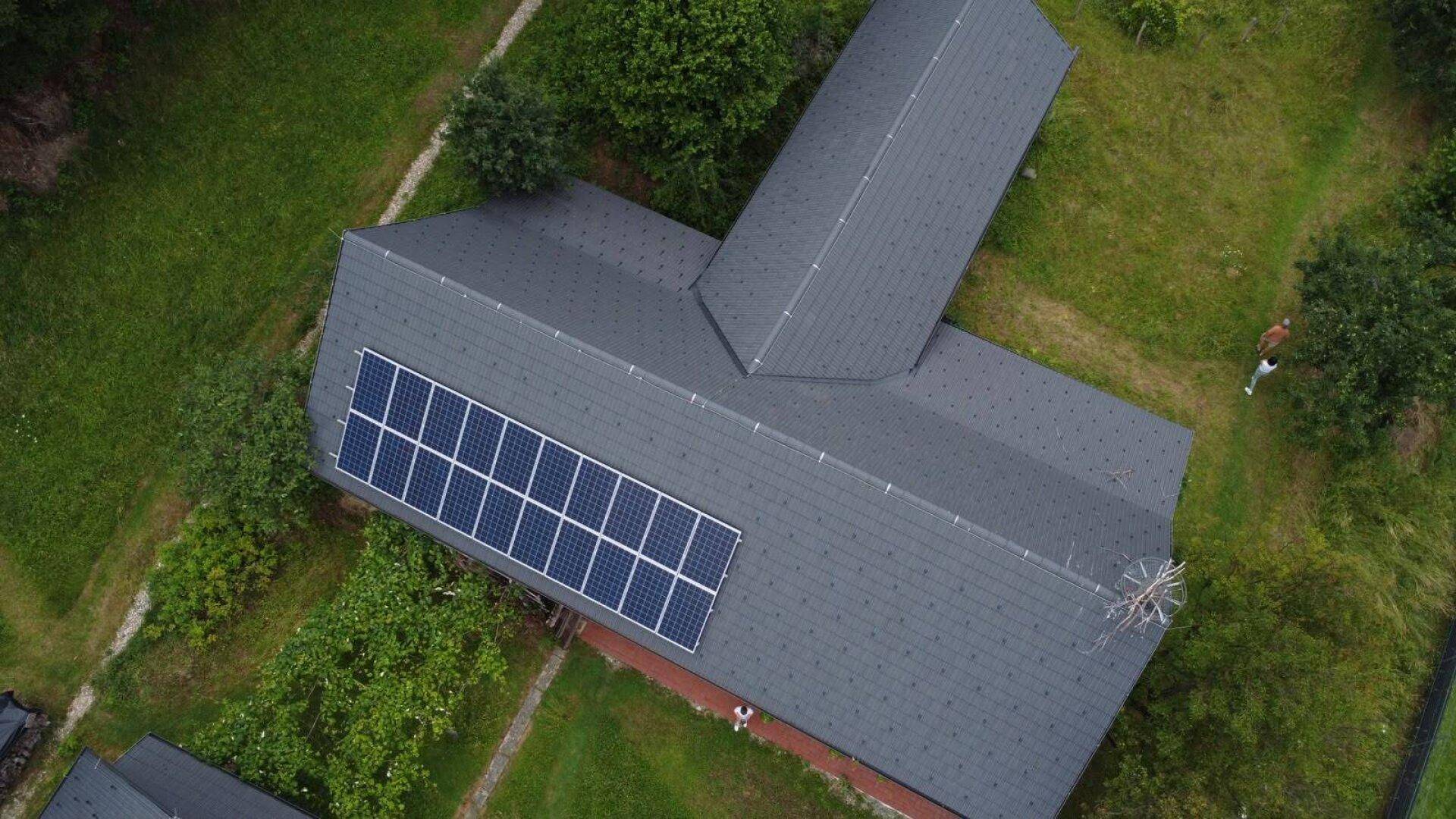 Nahaufnahme eines Daches mit Photovoltaikanlage und grauen Dachziegeln, umgeben von Bäumen.