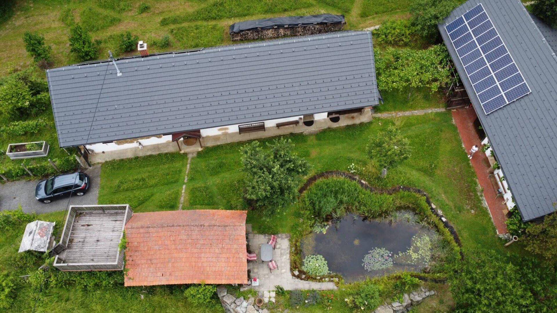 Überblick über ein ländliches Anwesen mit Gebäuden, einem Teich und parkenden Autos in grüner Umgebung.