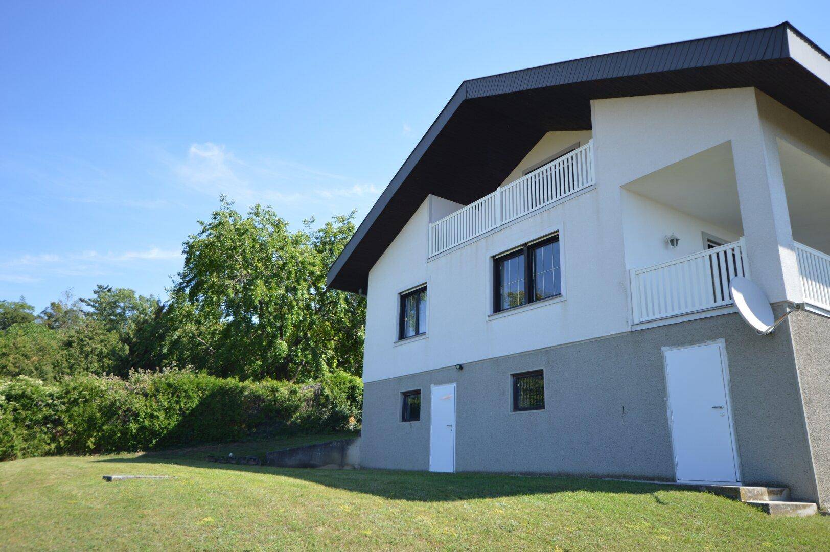 Seitenansicht des Hauses mit Balkonen und grüner Umgebung unter blauem Himmel.