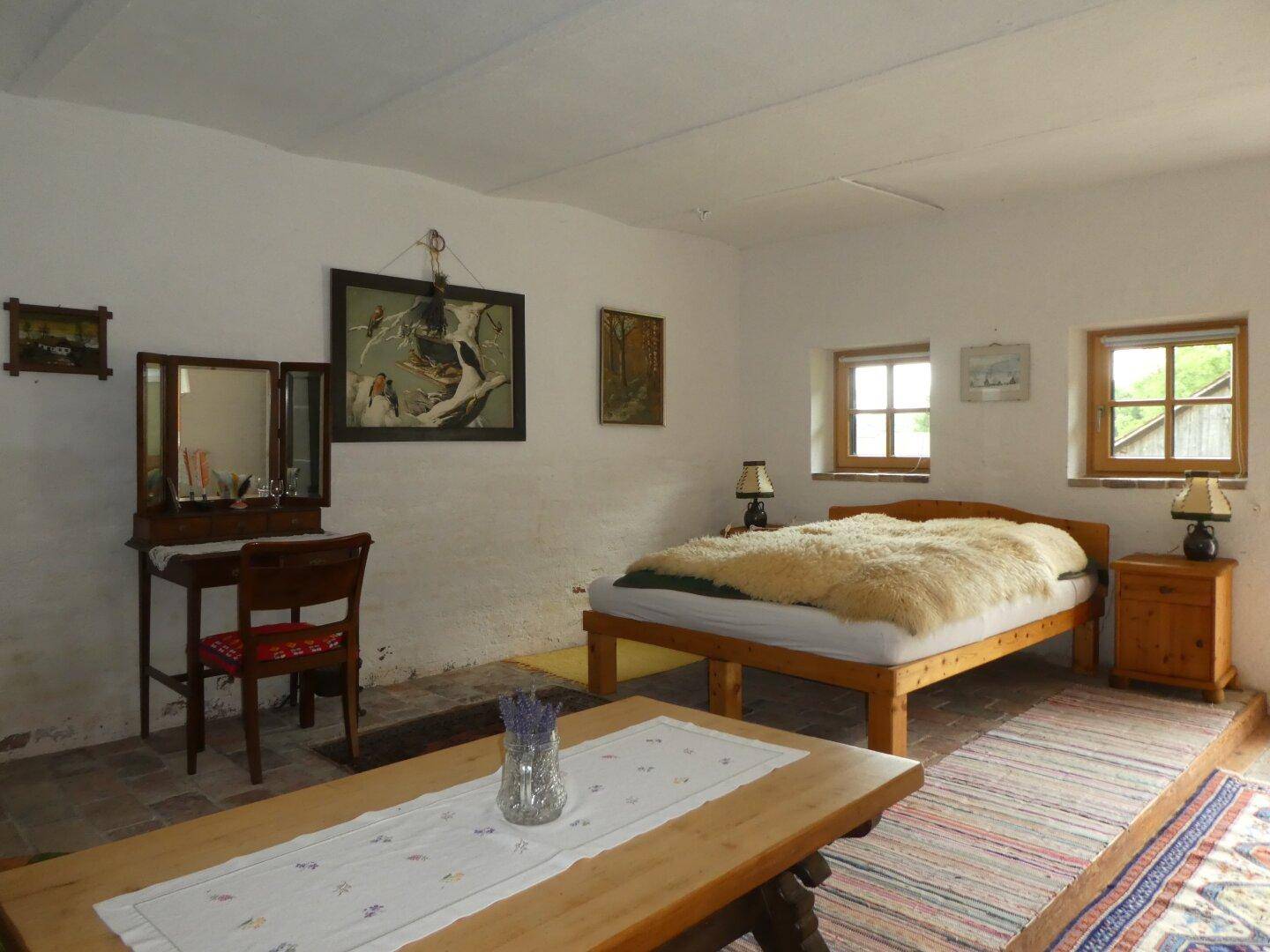Gemütliches Schlafzimmer im rustikalen Stil mit Holzbett, Schminktisch und Fliesenboden, hell durch zwei Fenster.