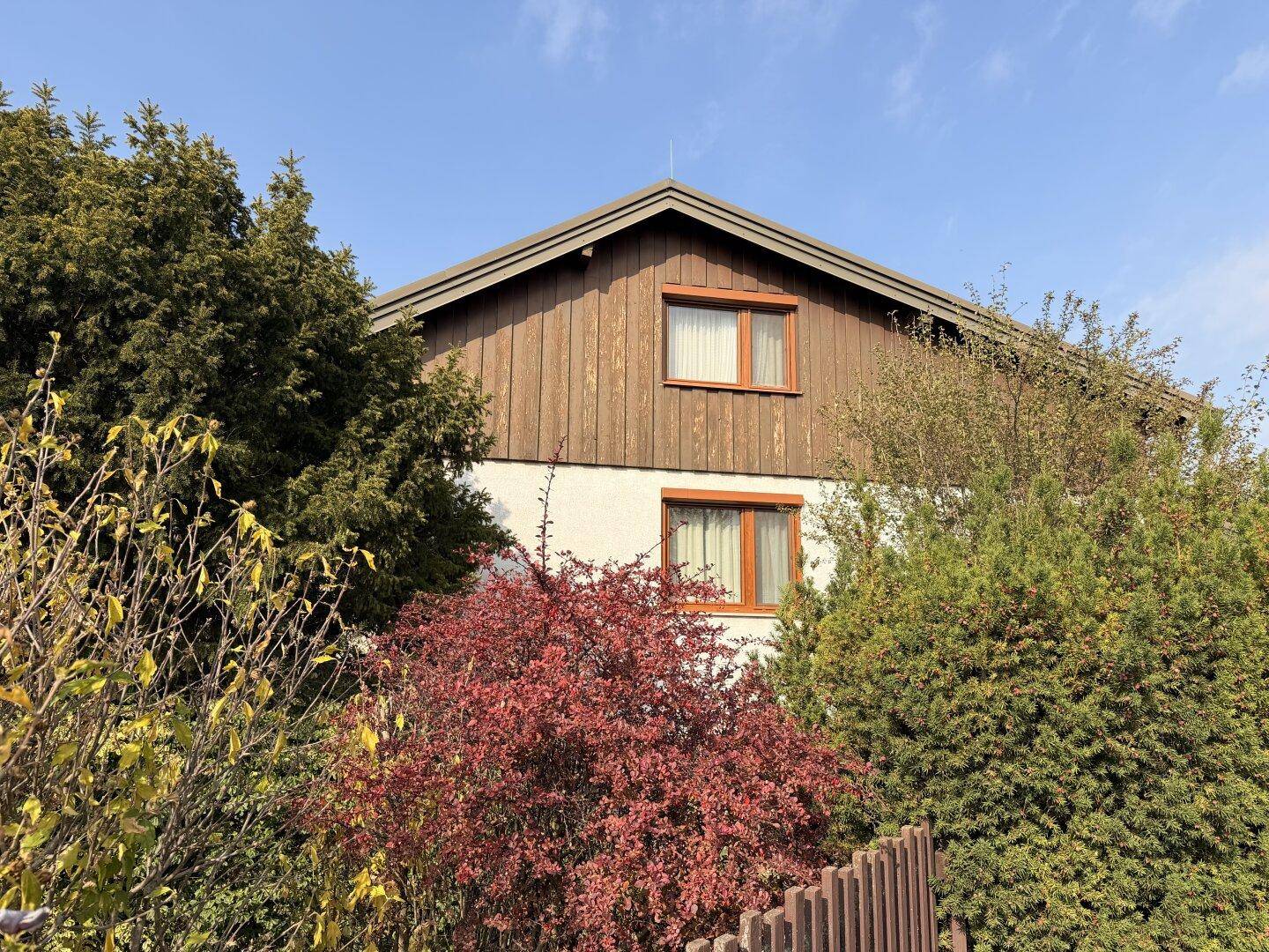 Ansicht des Hauses mit Holzverkleidung und üppiger Bepflanzung im Vorgarten unter blauem Himmel.