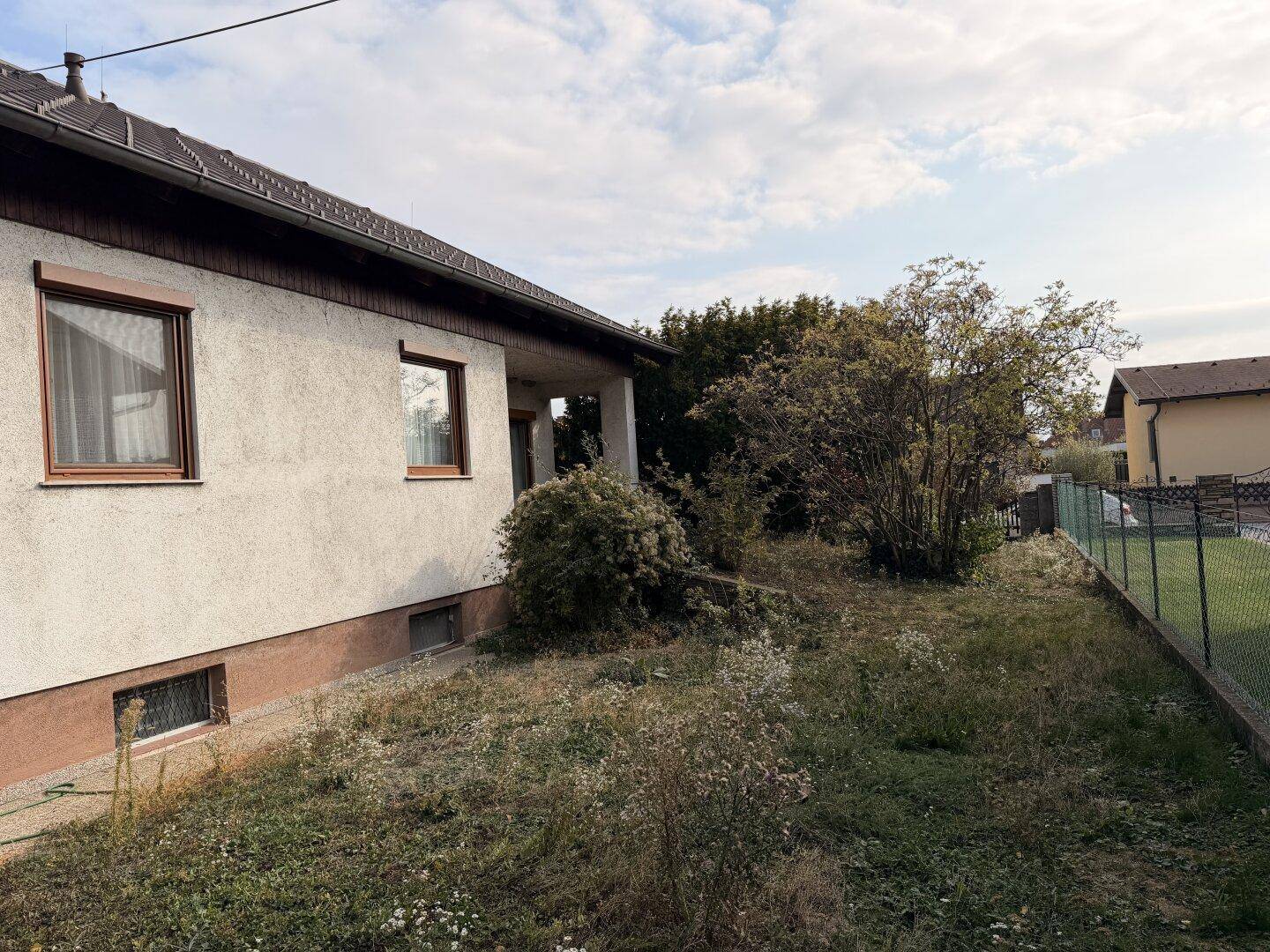 Ungepflegter Gartenbereich neben dem Haus mit Rasen, Büschen und einem Zaun.
