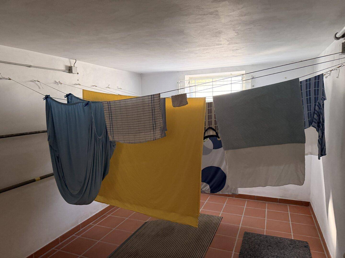 Trockenraum im Keller mit Wäscheleinen und Terrakotta-Fliesenboden, beleuchtet durch ein kleines Fenster.
