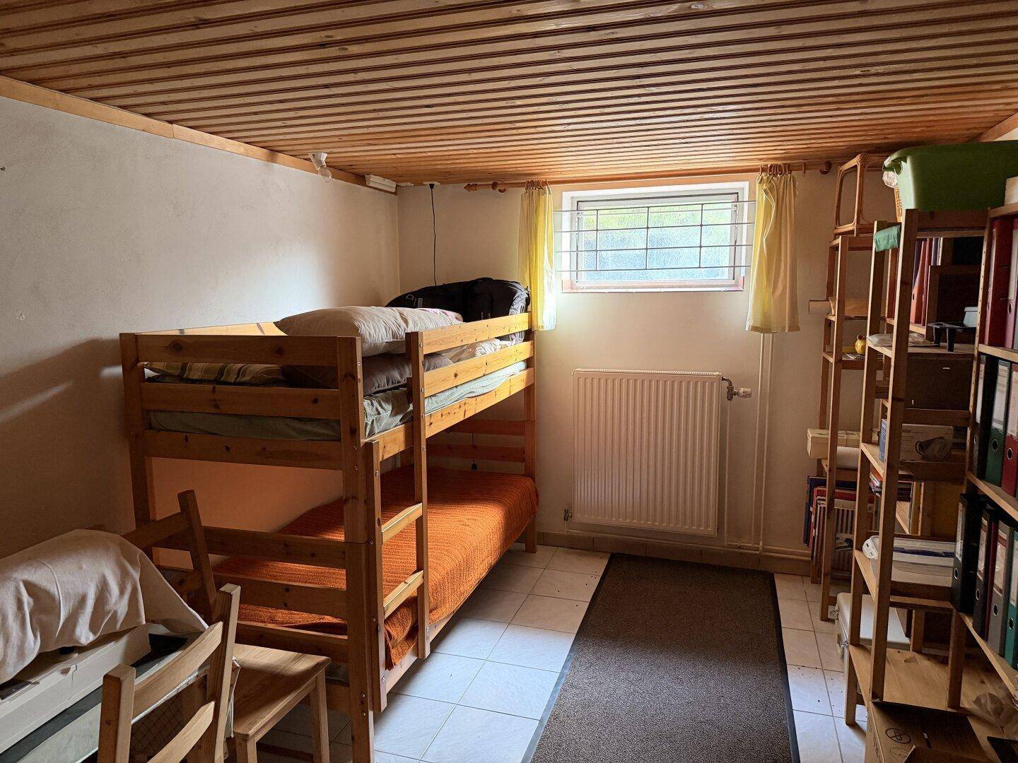 Kellerzimmer mit Etagenbett, Bücherregal und kleinem Fenster, ausgestattet mit Holzdecke.