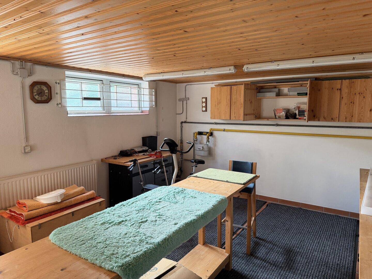 Hobbyraum im Keller mit Werkbank, Fitnessgerät und Holzdecke, beleuchtet durch ein Fenster.