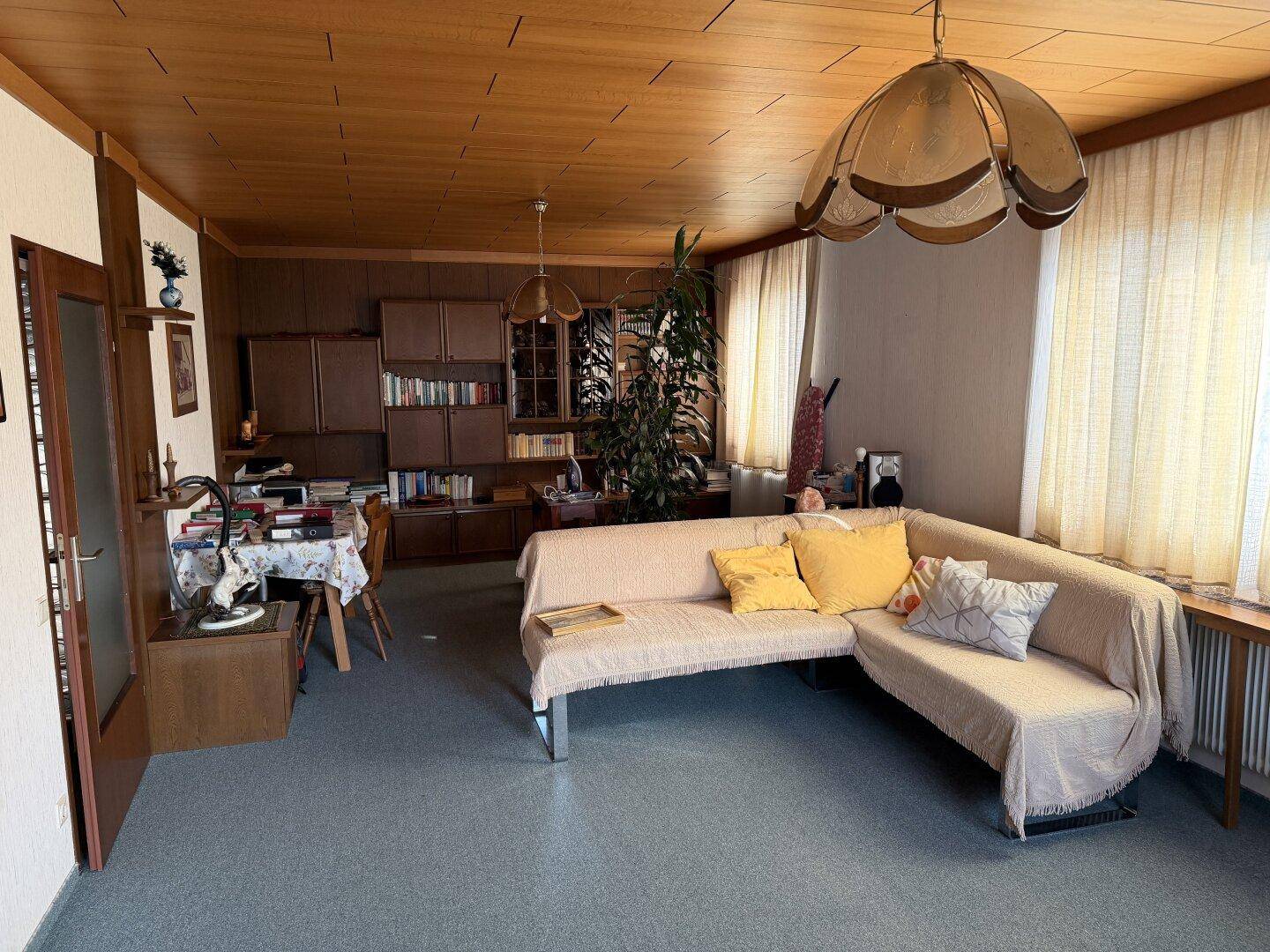 Wohnbereich mit Holzdecke, Teppichboden und traditionellen Möbeln, inklusive Bücherregal und Esstisch.