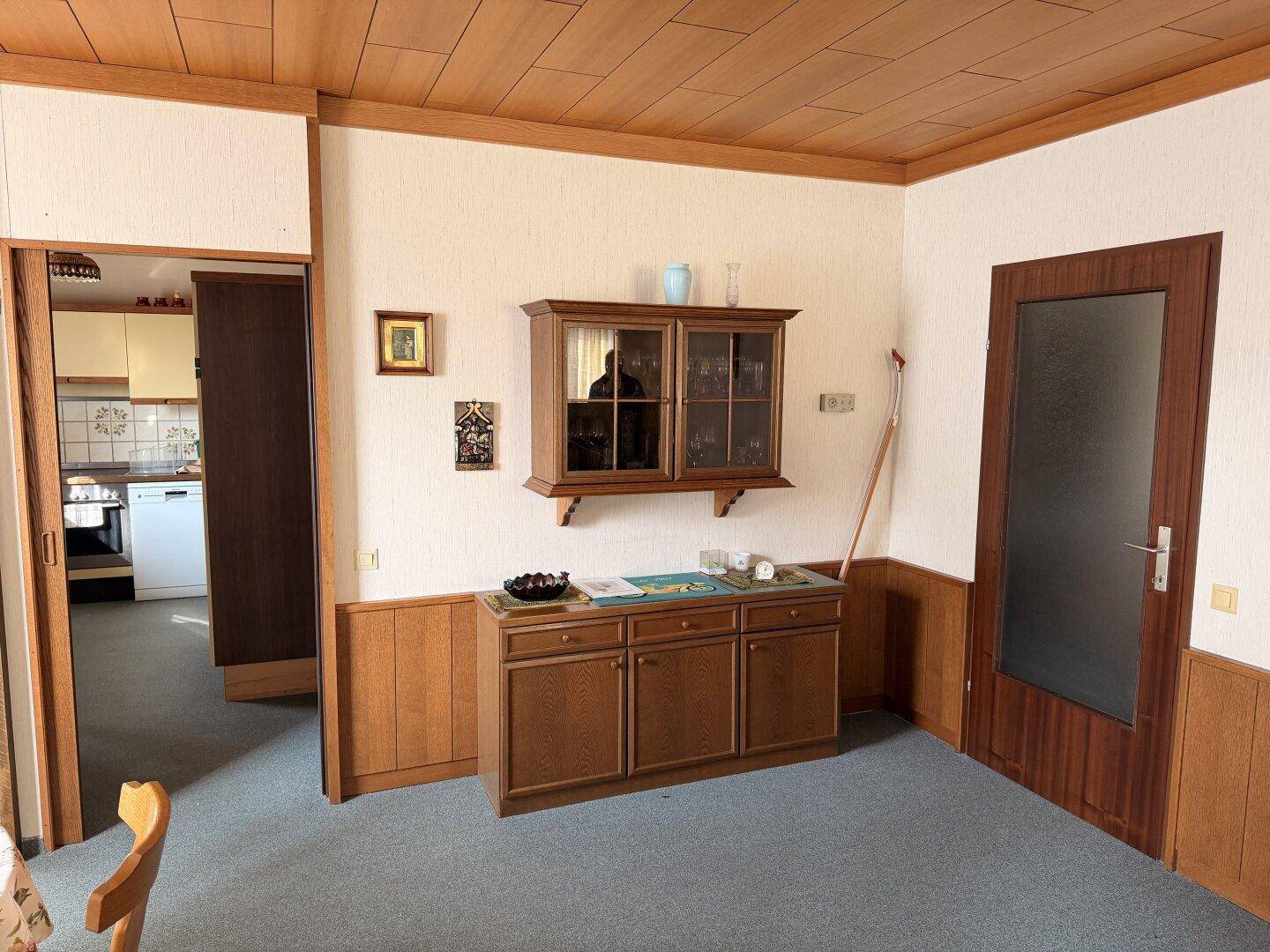 Essbereich mit Holzvertäfelung, Teppichboden und einem Durchgang zur Küche, ausgestattet mit einem Vitrinenschrank.