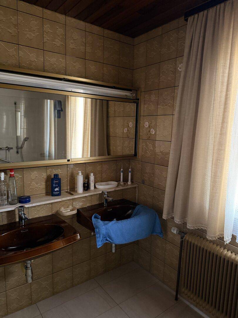 Badezimmer mit braunen Fliesen, zwei Waschbecken und einem großen Spiegel, ausgestattet mit einem Fenster.