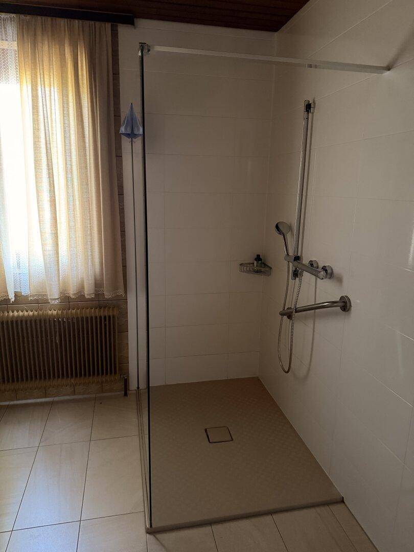 Modernisierte Dusche mit Glasabtrennung und hellen Fliesen, neben einem Fenster mit Vorhang.