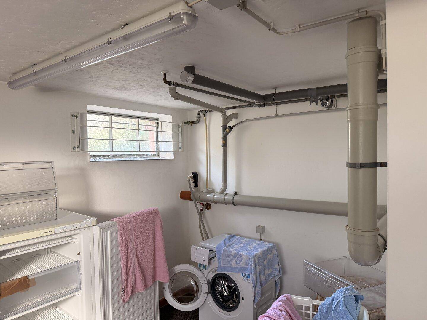 Waschküche im Keller mit Waschmaschine, Trockner und sichtbaren Rohren an der Decke.