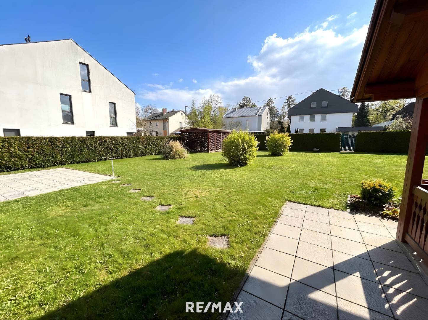 Sonniger Garten mit Rasenfläche, gepflasterter Terrasse und Hecke, angrenzend an ein modernes Gebäude.