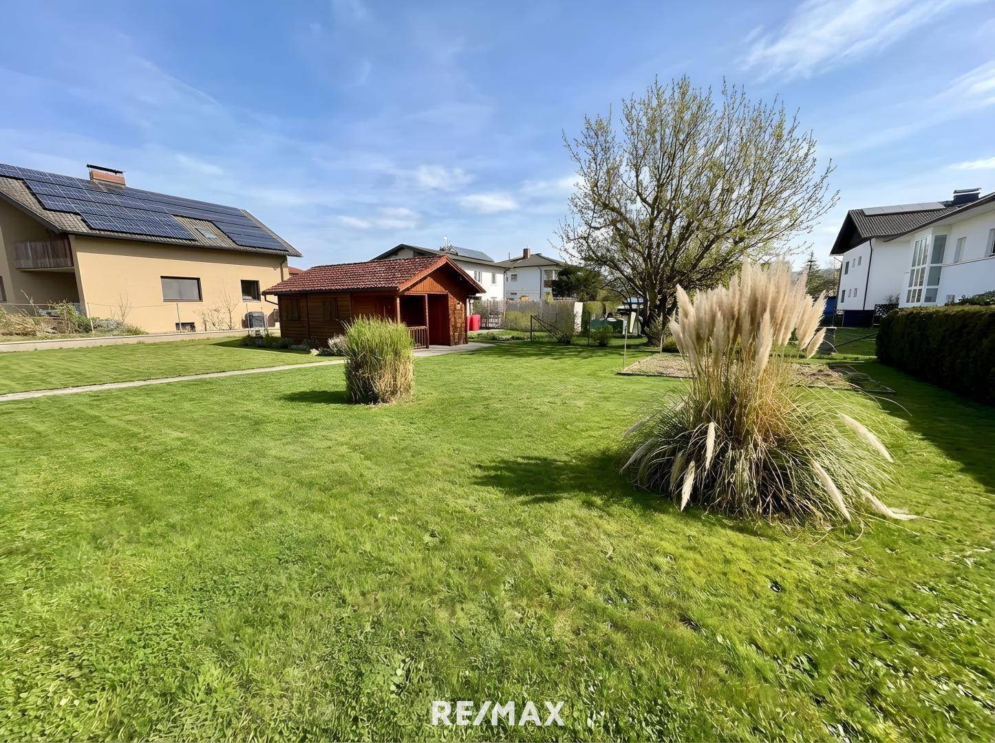 Weitläufiger Garten mit Rasen, Ziergräsern und einem kleinen Holzschuppen, bei sonnigem Wetter.