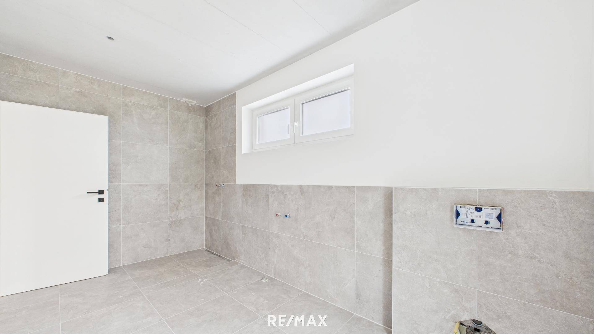 Geräumiges Badezimmer im Rohbau mit grauen Fliesen und Fenster für natürliche Belüftung und Licht.