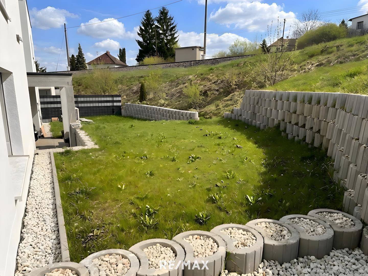 Hinterer Gartenbereich mit Rasenfläche, Stützmauern und dekorativen Steinelementen, umgeben von Grün.