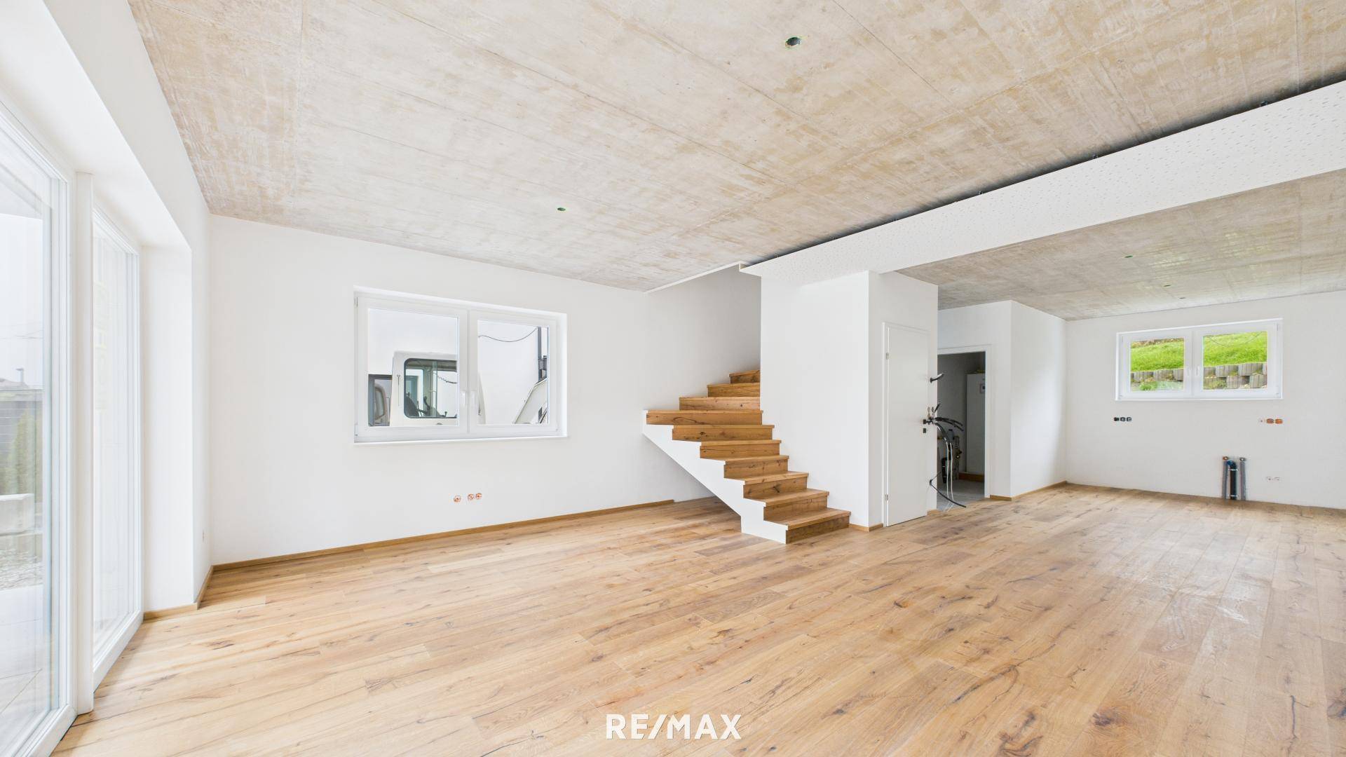 Geräumiger, heller Wohnbereich im Rohbau mit Holzboden, großen Fenstern und offener Treppe.