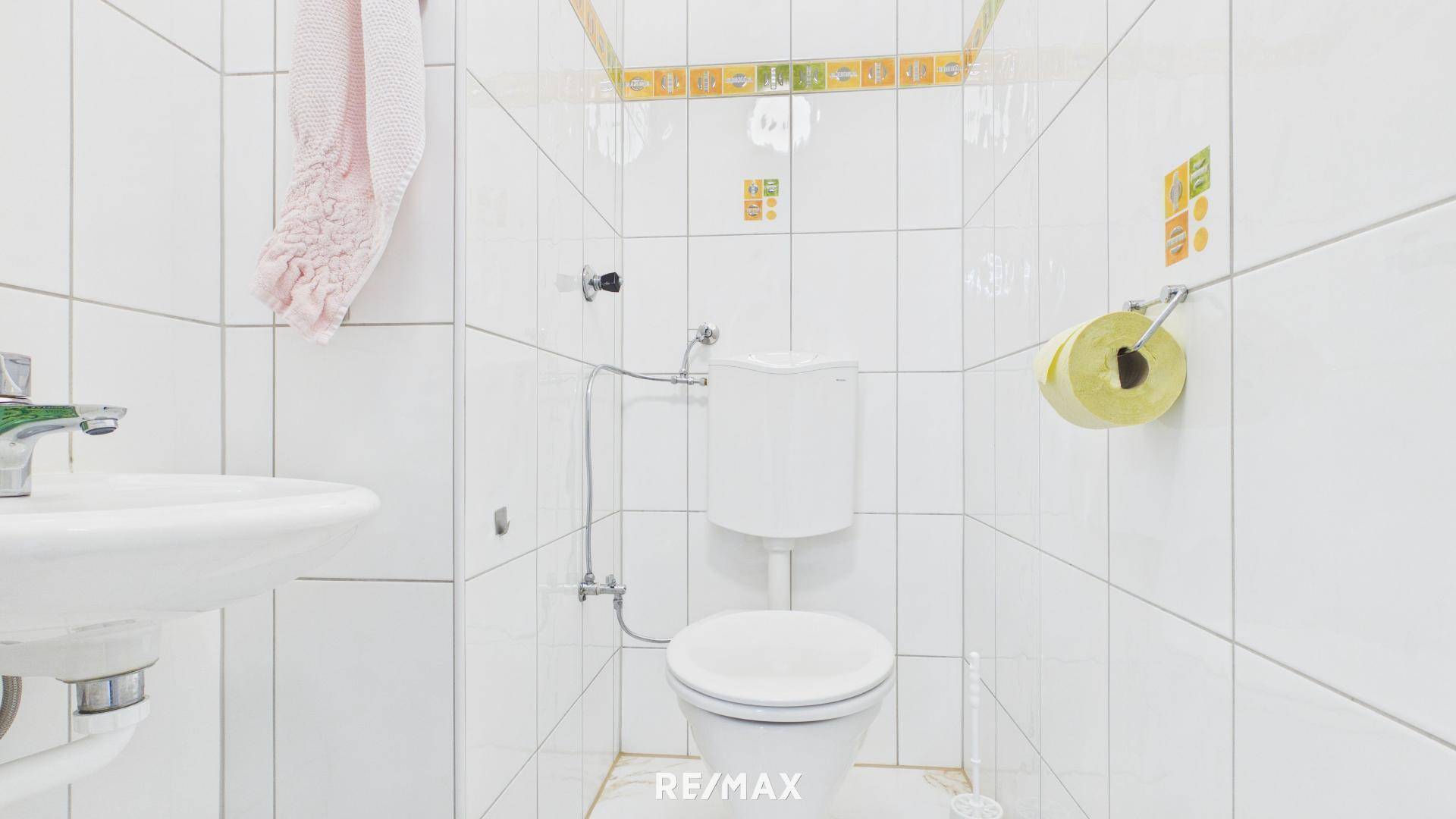 Separates WC mit weißer Toilette, kleinem Waschbecken und hellen Fliesen.
