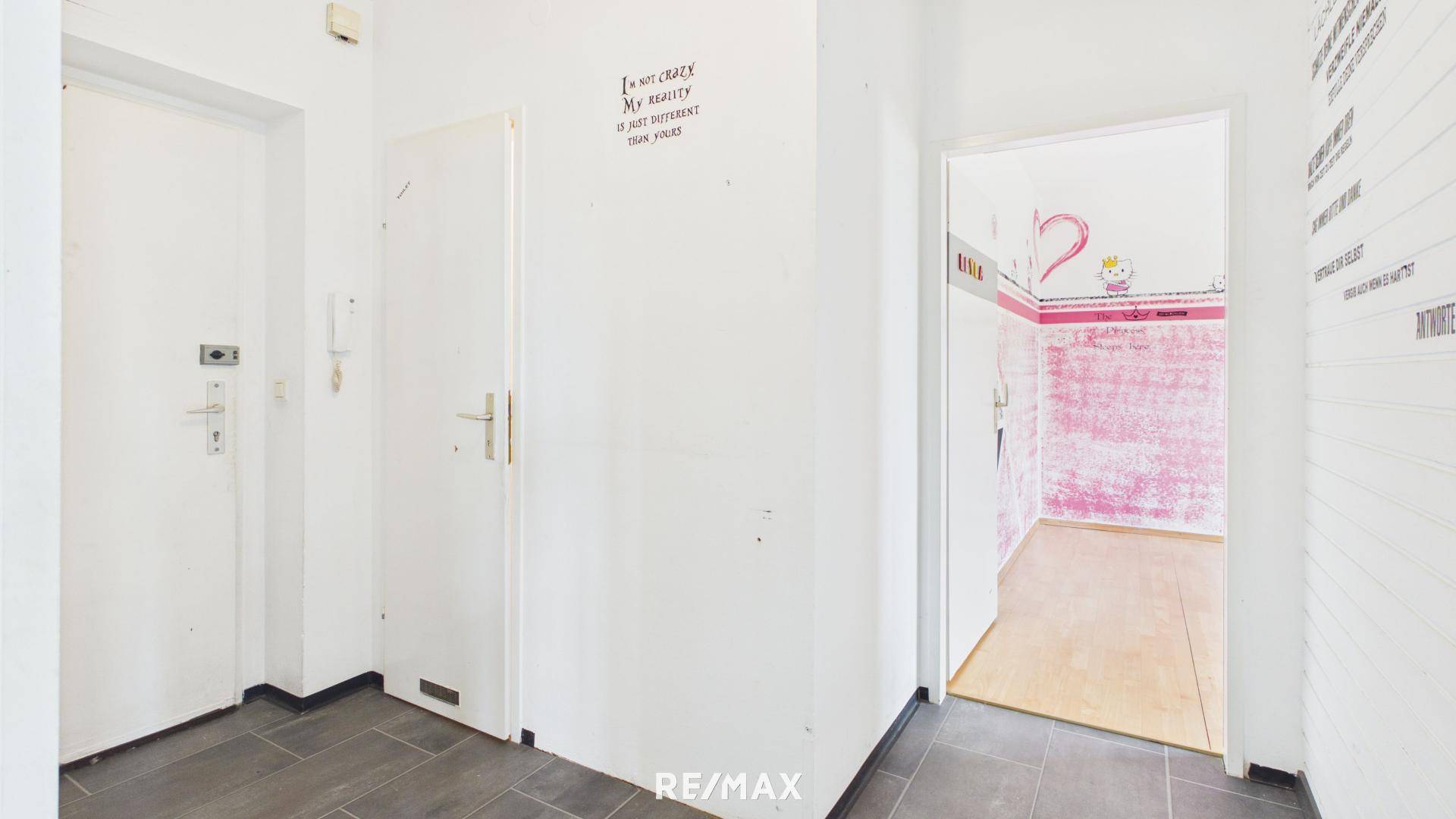 Eingangsbereich mit weißer Tür und grauem Fliesenboden, der zu einem Zimmer mit rosa Wänden führt.
