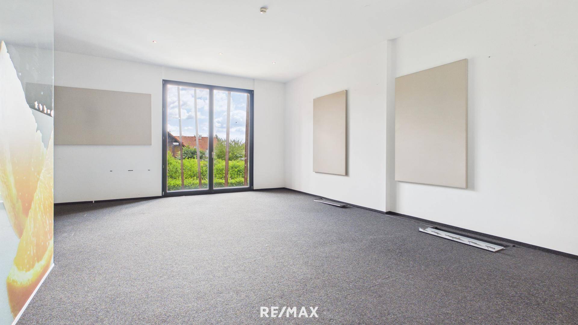 Leeres Bürozimmer mit dunklem Teppichboden und großer Glas-Schiebetür mit Blick ins Grüne.