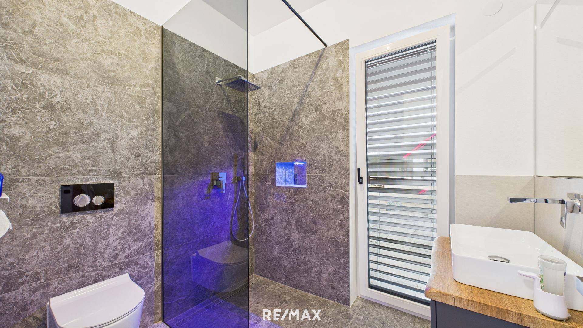 Modernes Badezimmer mit grauen Fliesen, begehbarer Dusche und wandhängendem WC, beleuchteter Nische.