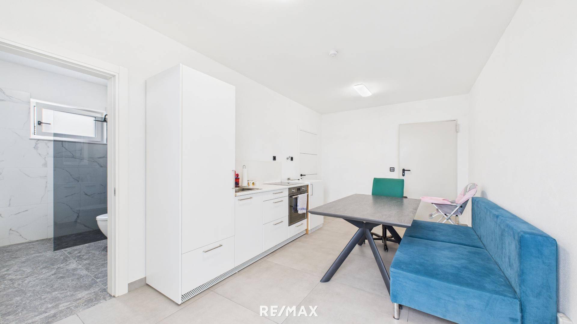 Kleine Küchenzeile mit weißen Schränken, Esstisch und blauem Sofa, angrenzend an ein Badezimmer.