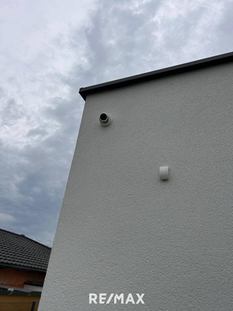 Moderne Außenfassade eines Neubaus mit Überwachungskamera und Flachdach unter bewölktem Himmel.