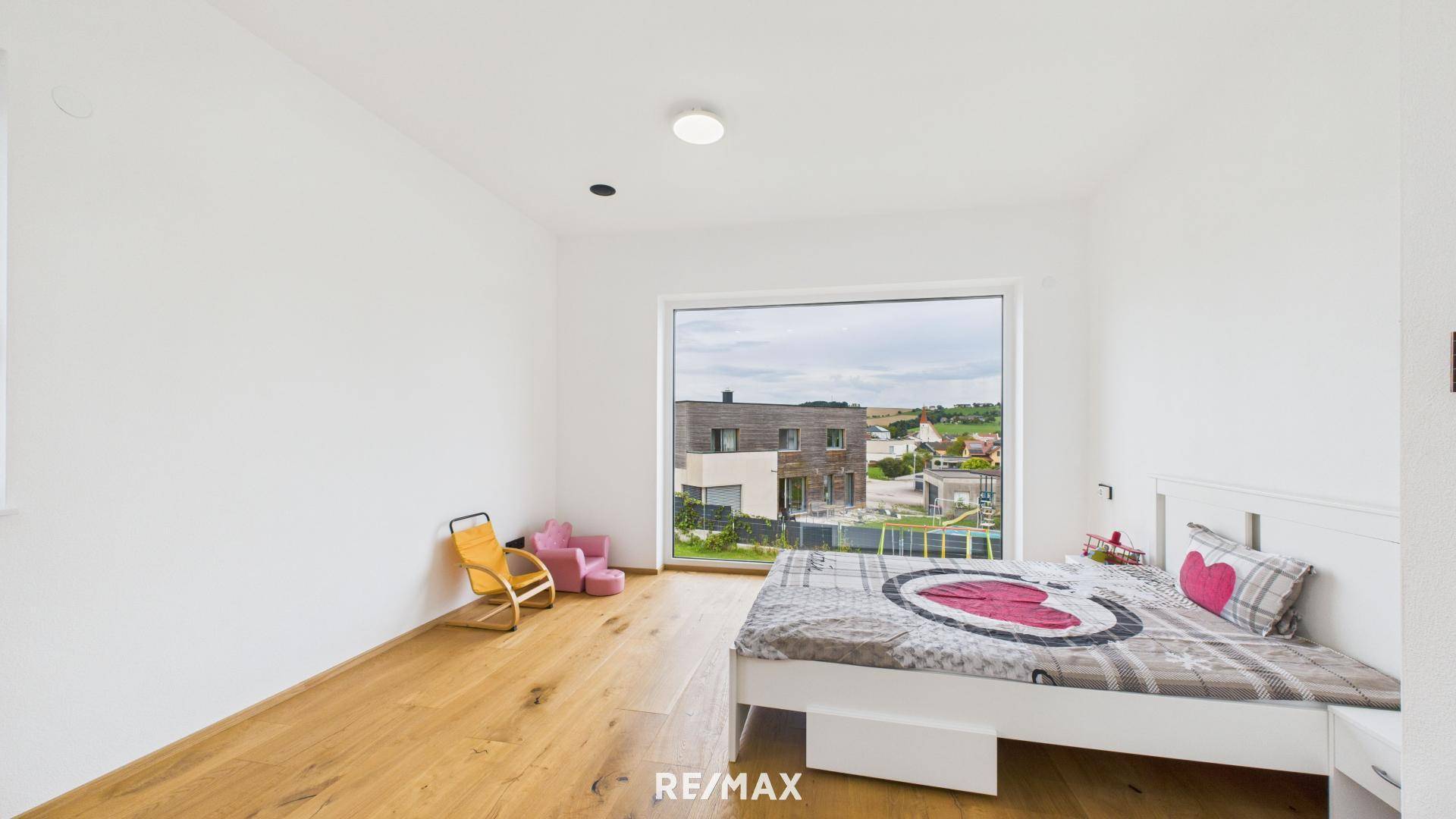 Helles Kinderzimmer mit Holzboden, Bett und großem Fenster mit Blick auf die Nachbarschaft.