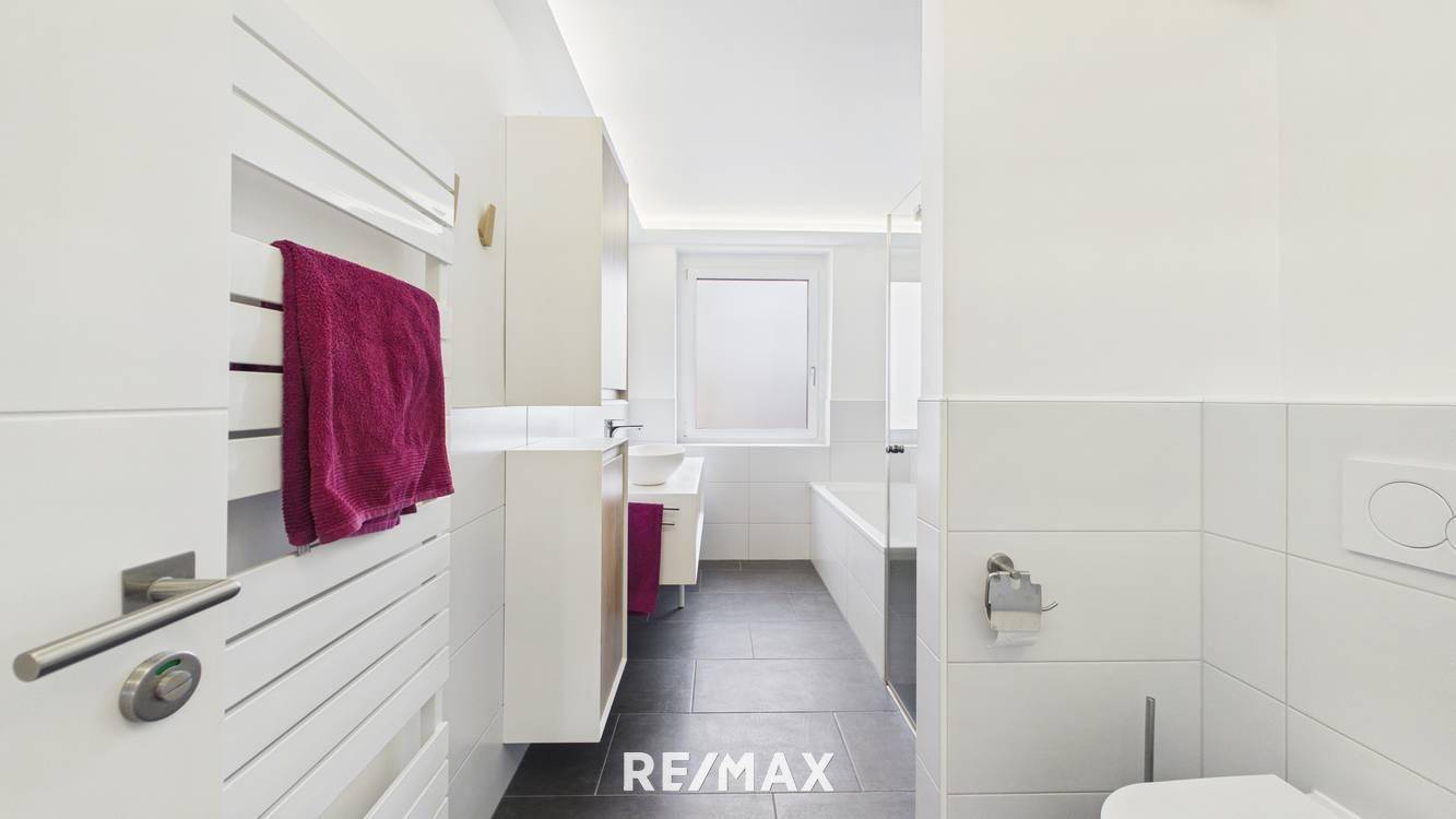 Modernes Badezimmer mit Badewanne, dunklen Bodenfliesen, weißer Wandverkleidung und Fenster.