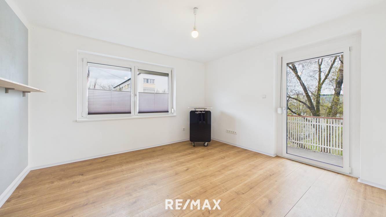 Helles Zimmer mit Holzboden, weiß gestrichenen Wänden und Zugang zu einem Balkon.