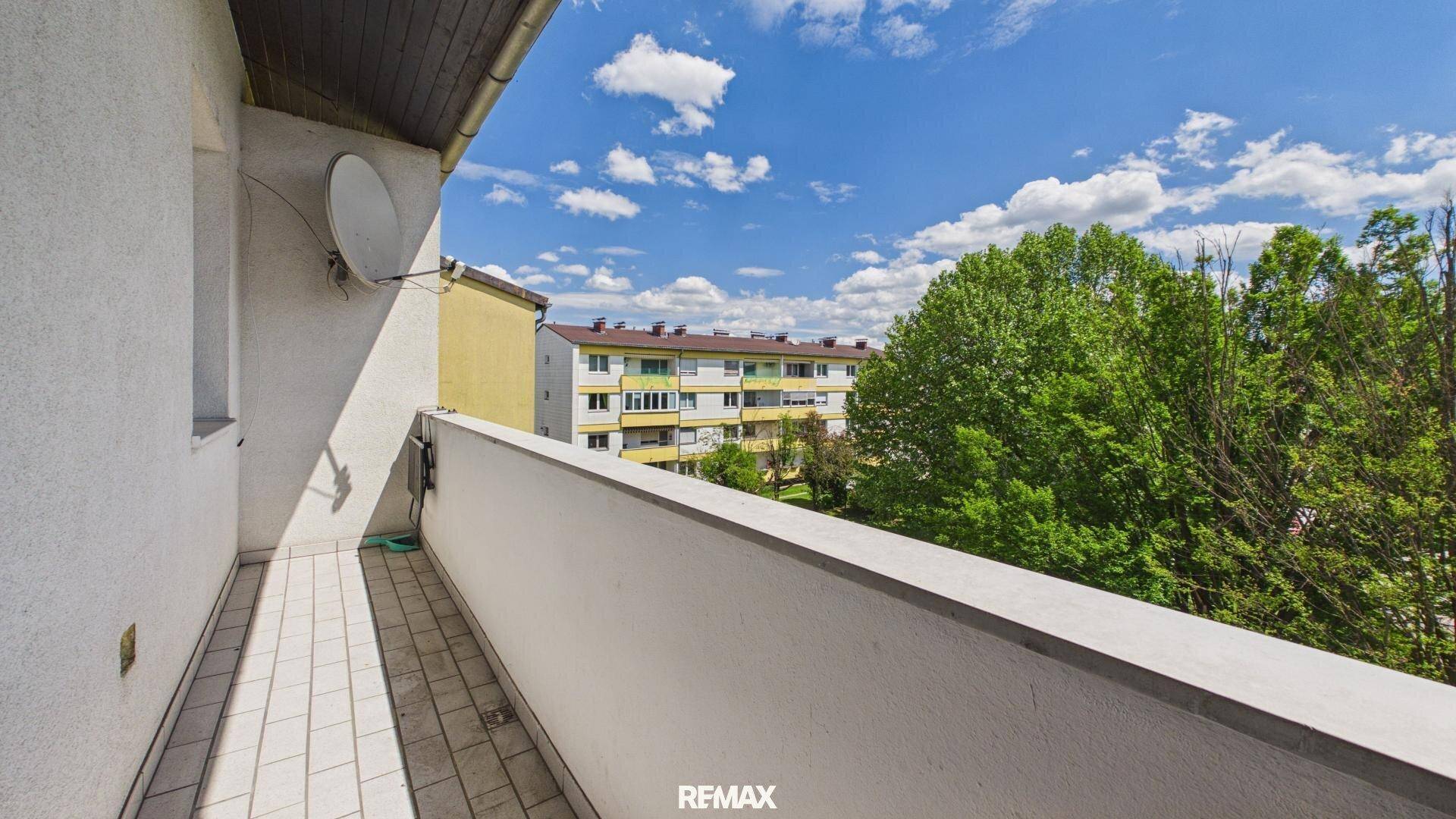 Heller Balkon mit Fliesenboden und Ausblick auf umliegende Gebäude und grüne Bäume.