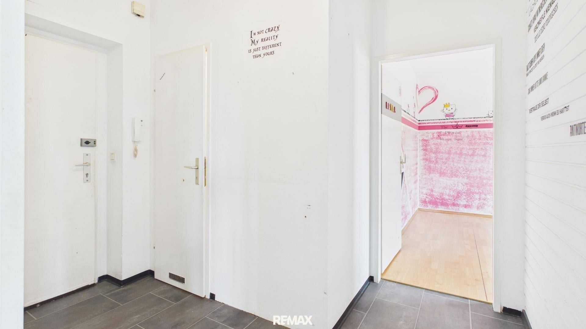 Eingangsbereich mit weiß gestrichenen Wänden, Sicherheitstür und Zugang zu einem Zimmer mit rosa Wänden.