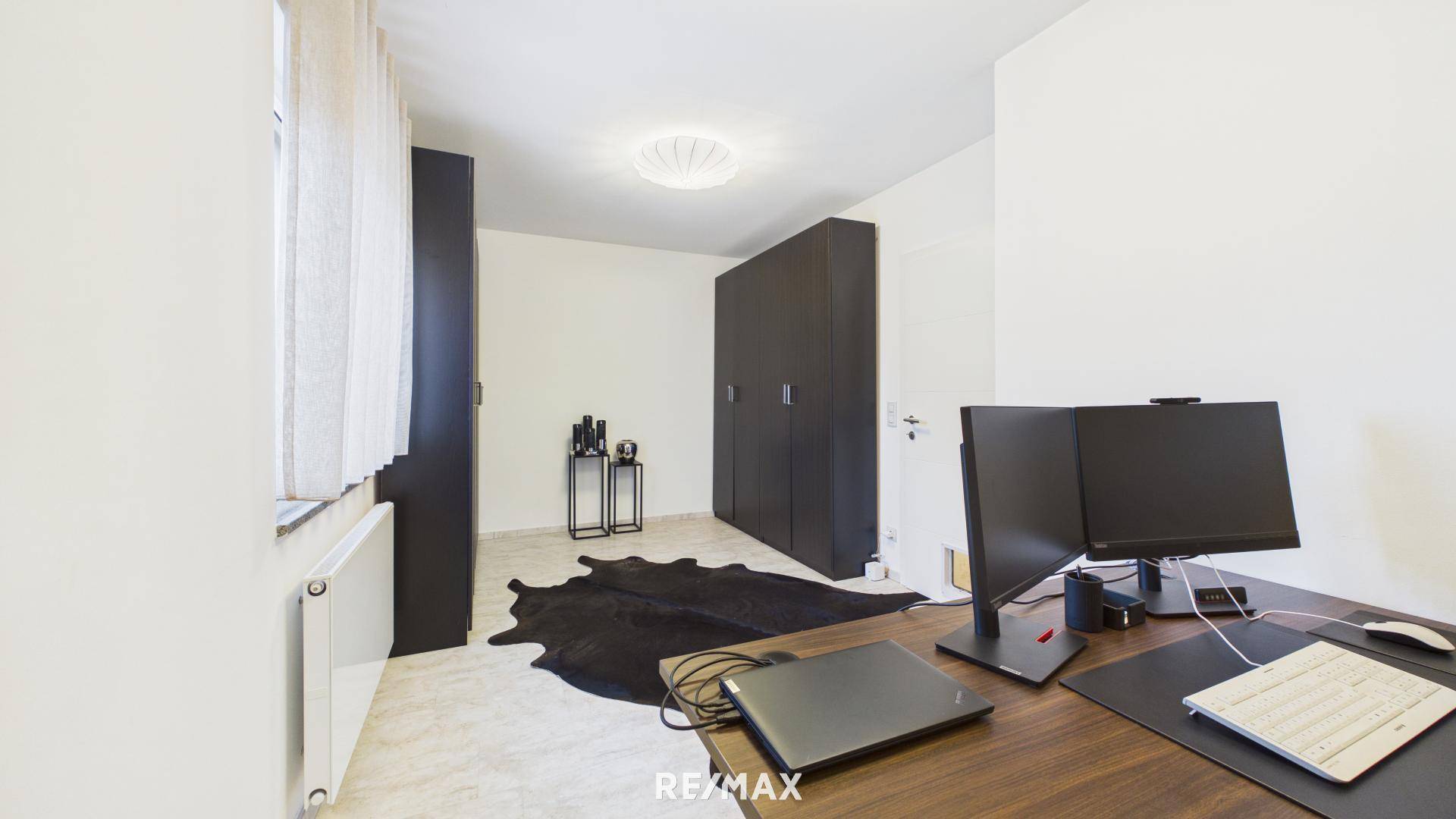 Helles Arbeitszimmer mit Schreibtisch, Computermonitoren und dunklen Schränken.