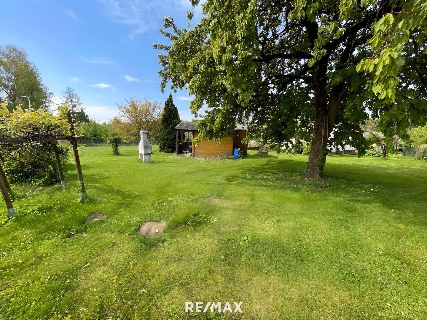 Großer Gartenbereich mit altem Baum, Gartenhaus und Grillplatz unter blauem Himmel.