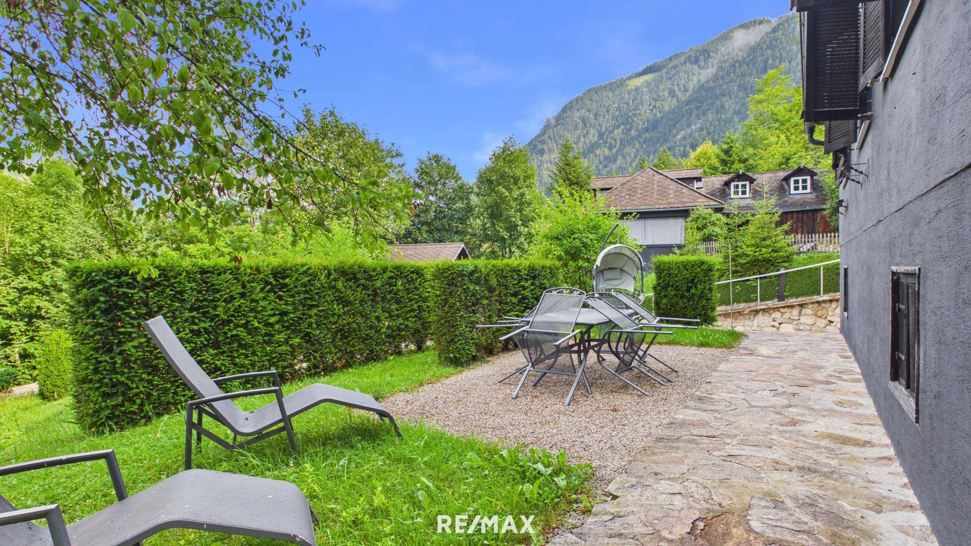 Terrasse mit Esstisch und Liegestühlen, umgeben von Hecken und Bergblick.