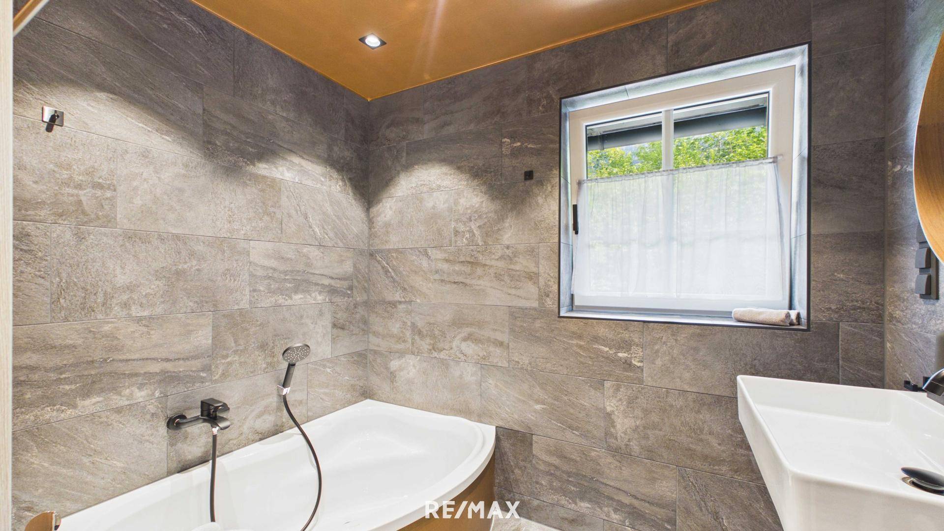Helles Badezimmer mit Badewanne, Fenster und modernen grauen Fliesen.