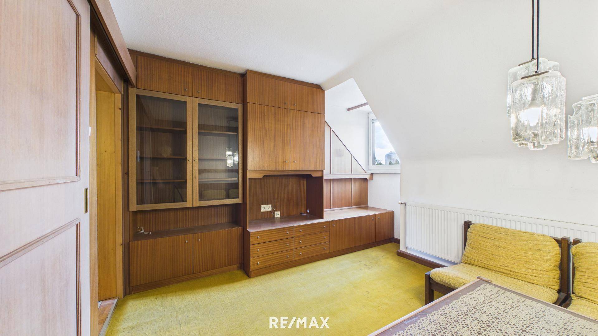 Wohnzimmer mit Einbauschränken aus Holz, gelbem Teppich und Dachschräge.