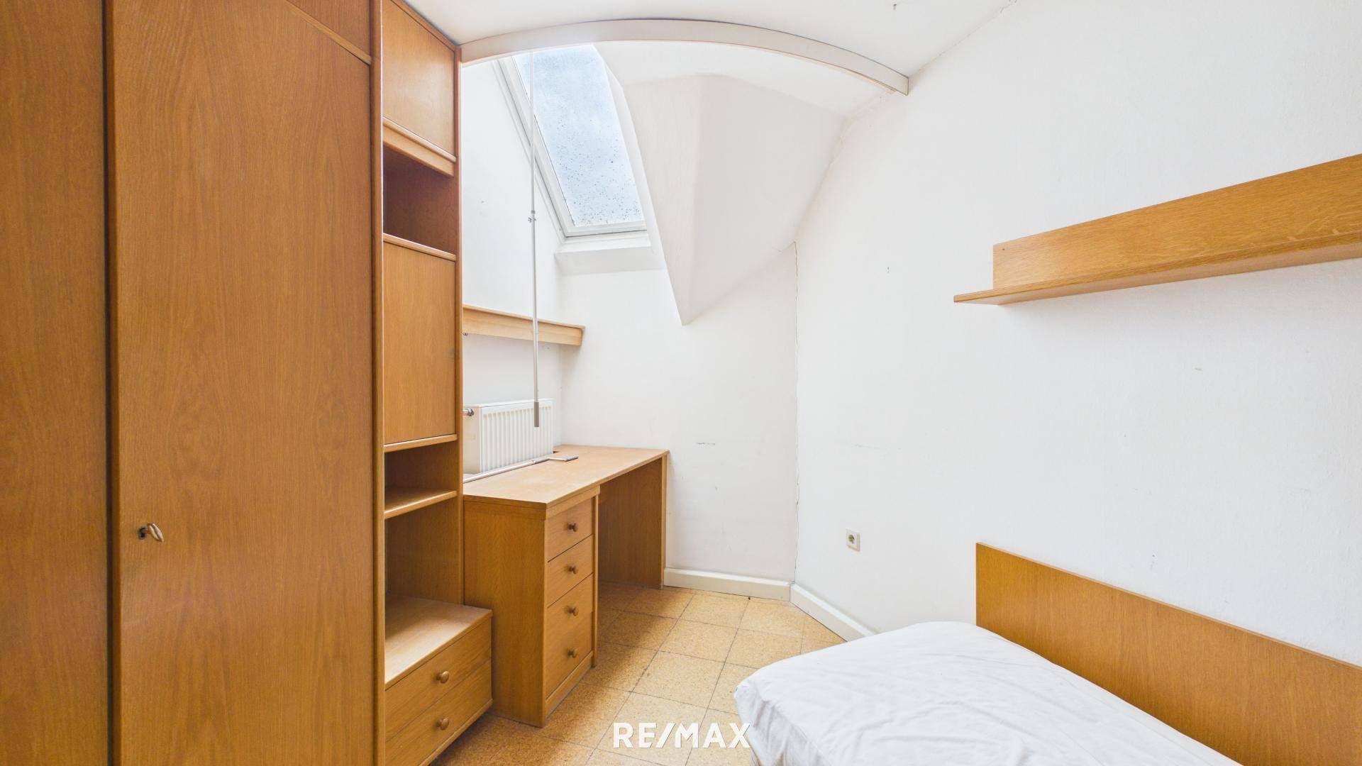 Kleines Zimmer mit Holzboden, Dachfenster, Schreibtisch und Bettgestell.