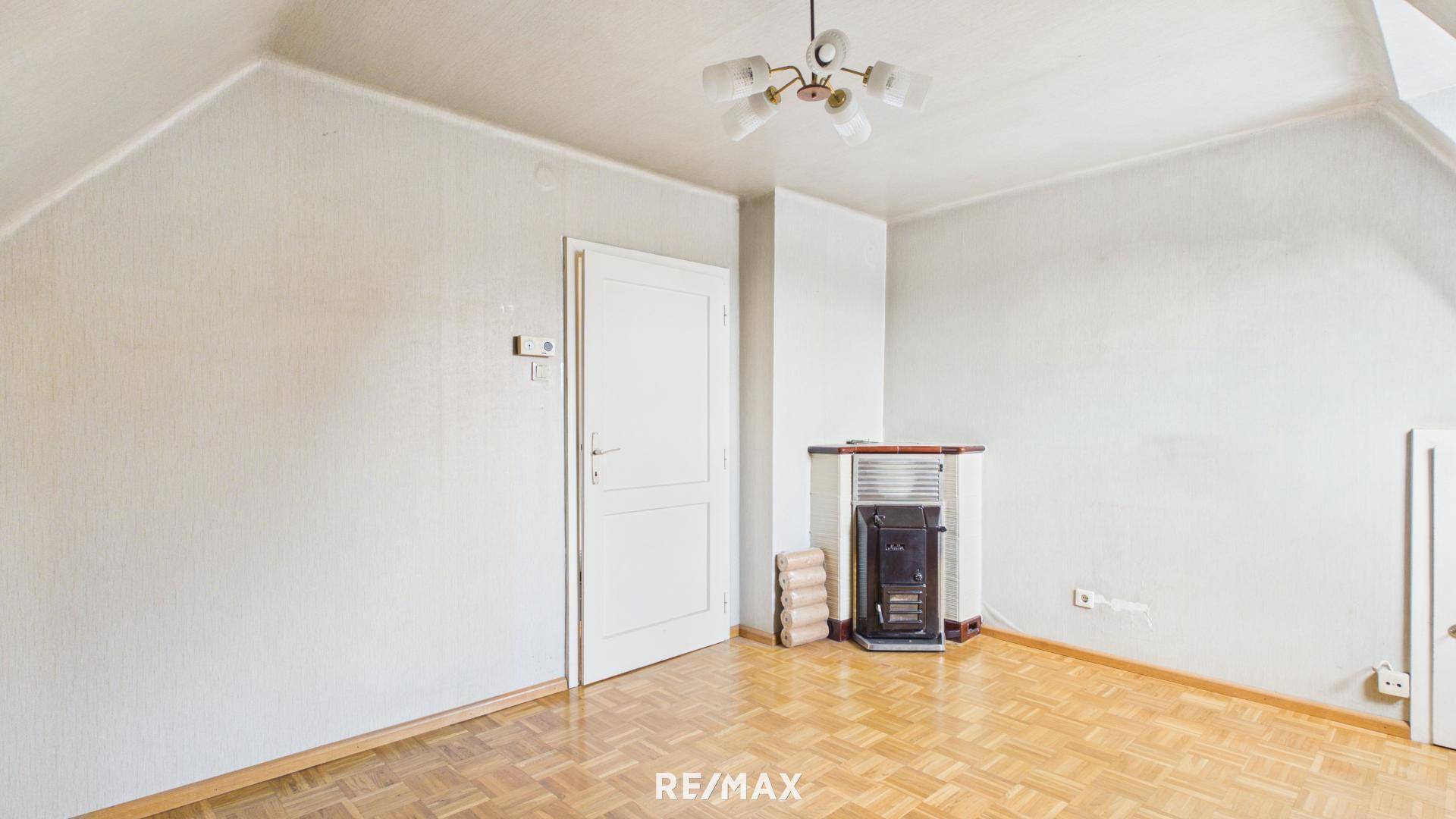 Helles Zimmer im Dachgeschoss mit Parkettboden und einem Ofen als Heizquelle, bereit zur Renovierung.