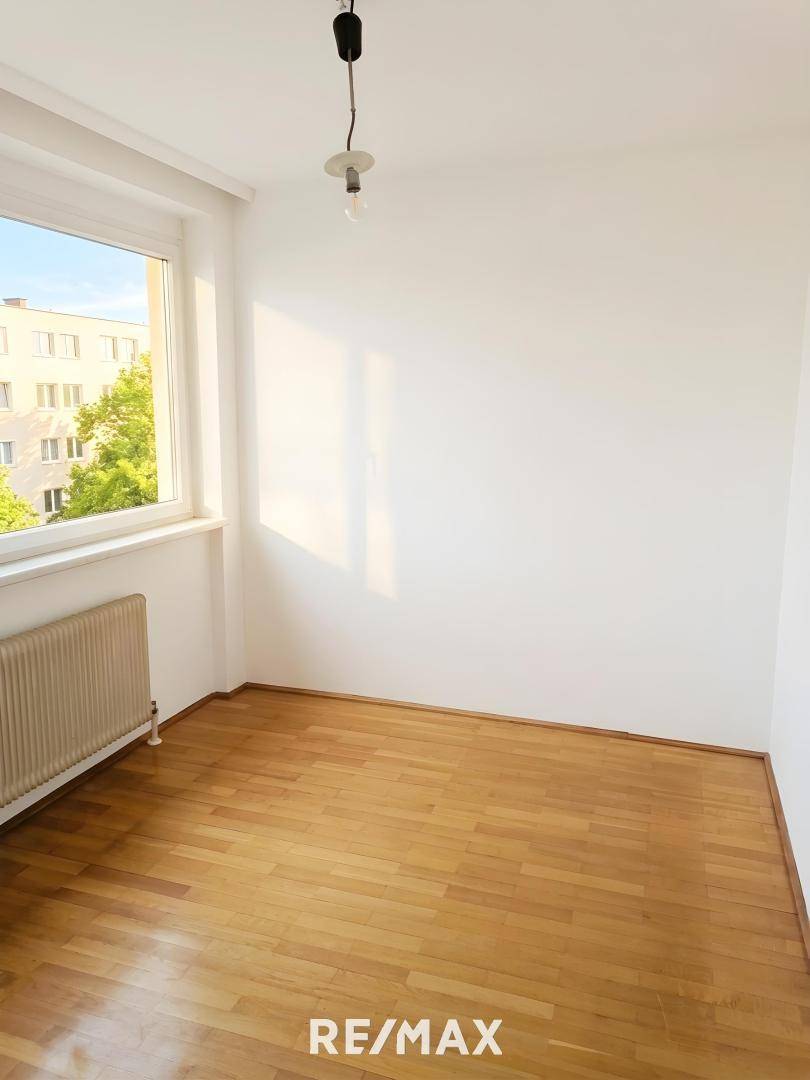 Sonniger Raum mit Holzboden und einem Fenster, das viel Tageslicht hereinlässt und eine grüne Aussicht bietet.