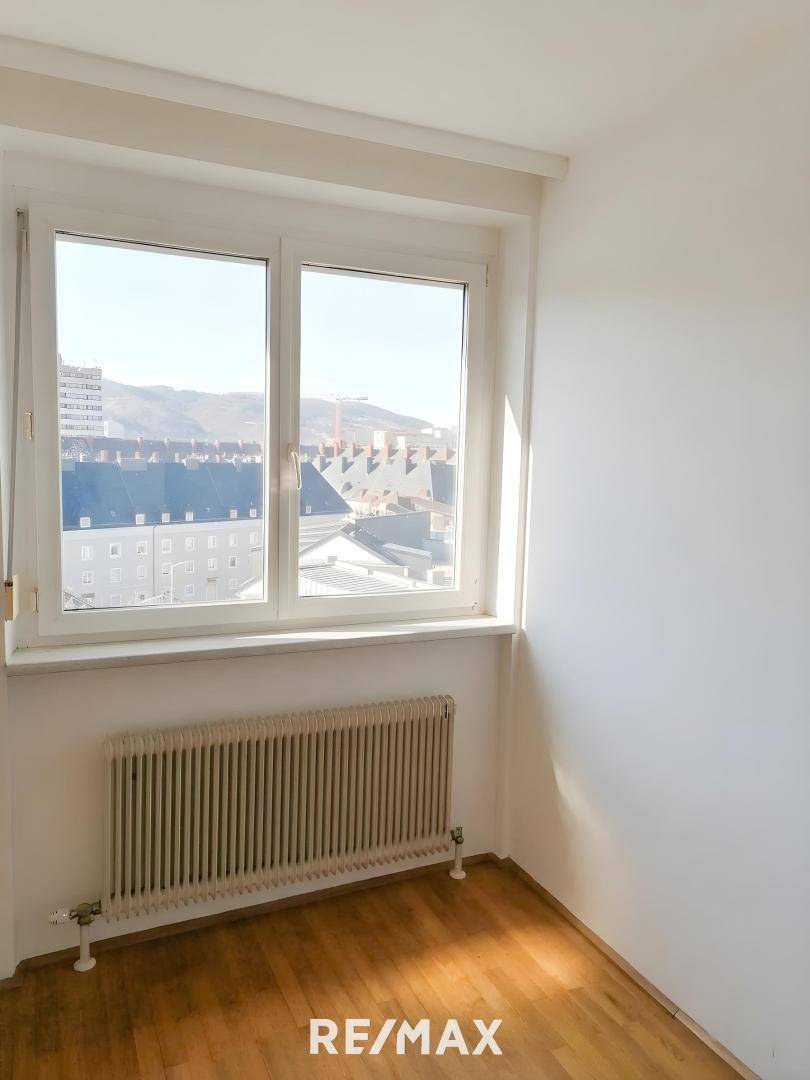 Heller Raum mit Parkettboden und einem Fenster mit Blick auf die Dächer der umliegenden Gebäude.