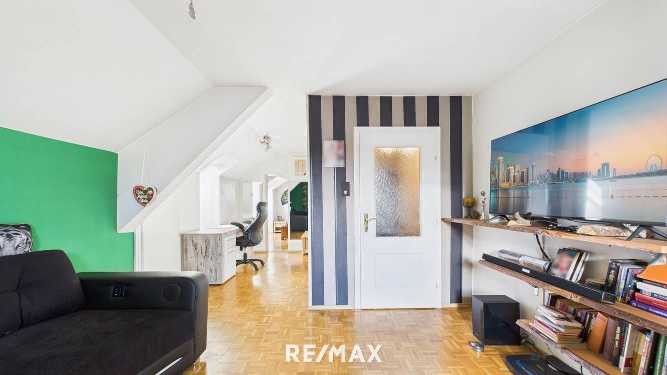 Gemütliches Wohnzimmer mit schwarzem Sofa, Fernseher und Blick in den Arbeitsbereich unter der Dachschräge.