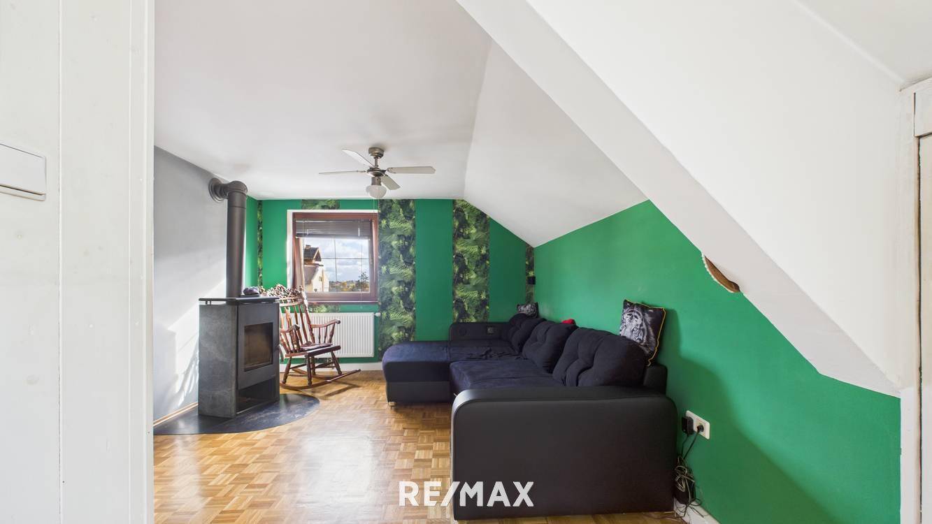 Geräumiges Wohnzimmer mit schwarzem Ecksofa, grünen Wänden und einem Kaminofen unter der Dachschräge.