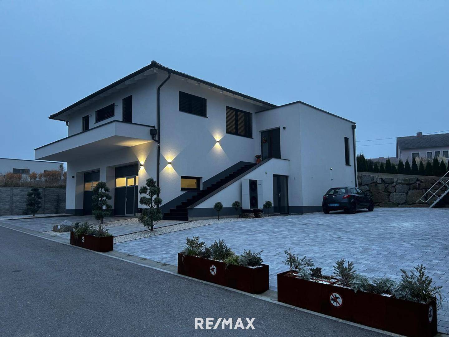 Modernes Haus bei Dämmerung mit beleuchteter Fassade, gepflasterter Auffahrt und dekorativen Pflanzen.
