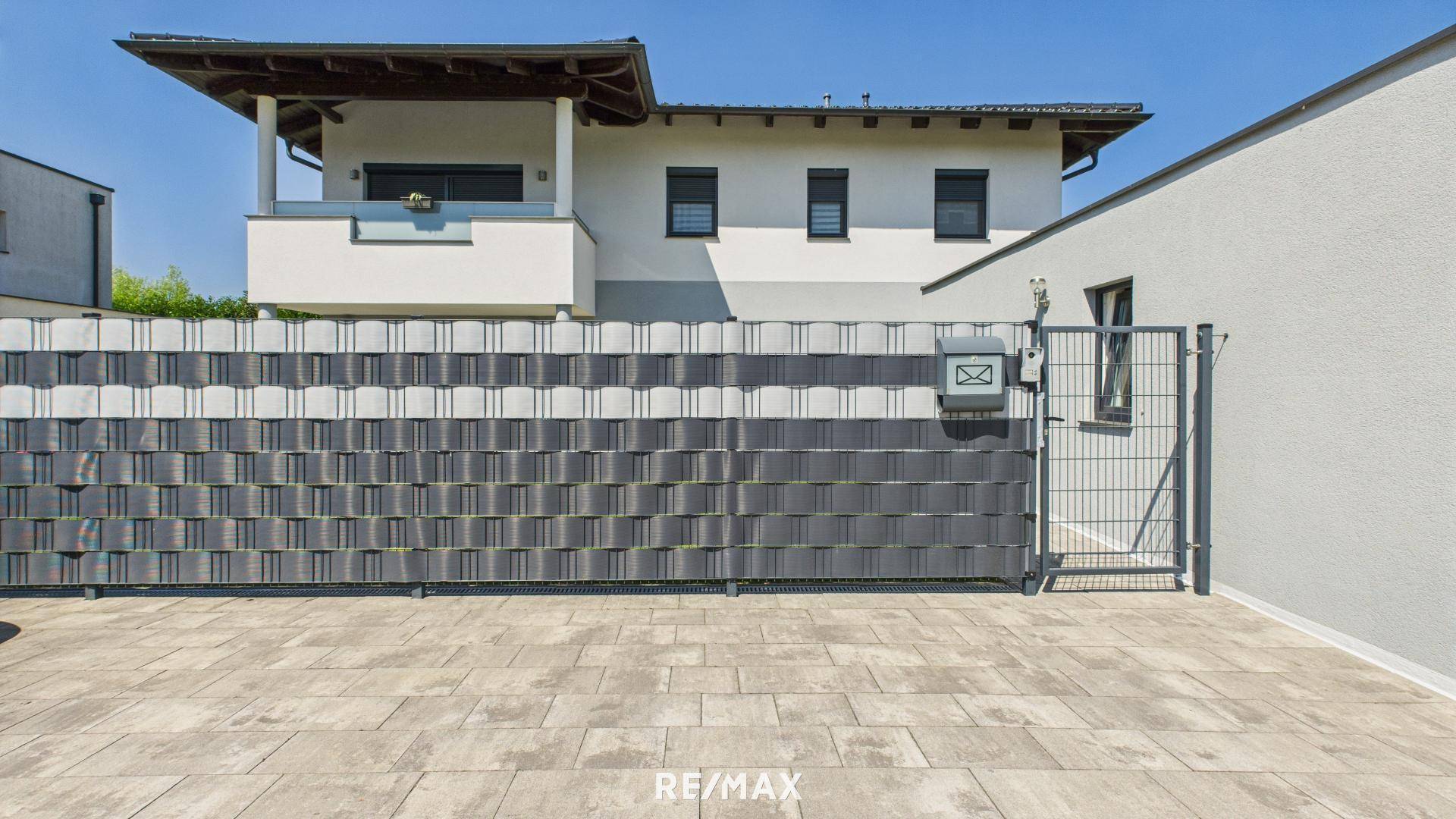 Moderne Hausfassade mit gepflastertem Vorplatz und blickdichtem Zaun.