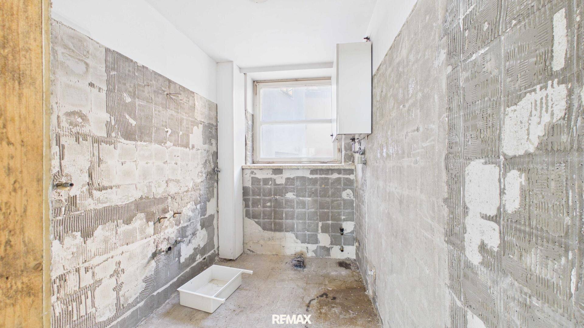Badezimmer im Rohbau mit entfernten Fliesen, Fenster und sichtbaren Installationen.