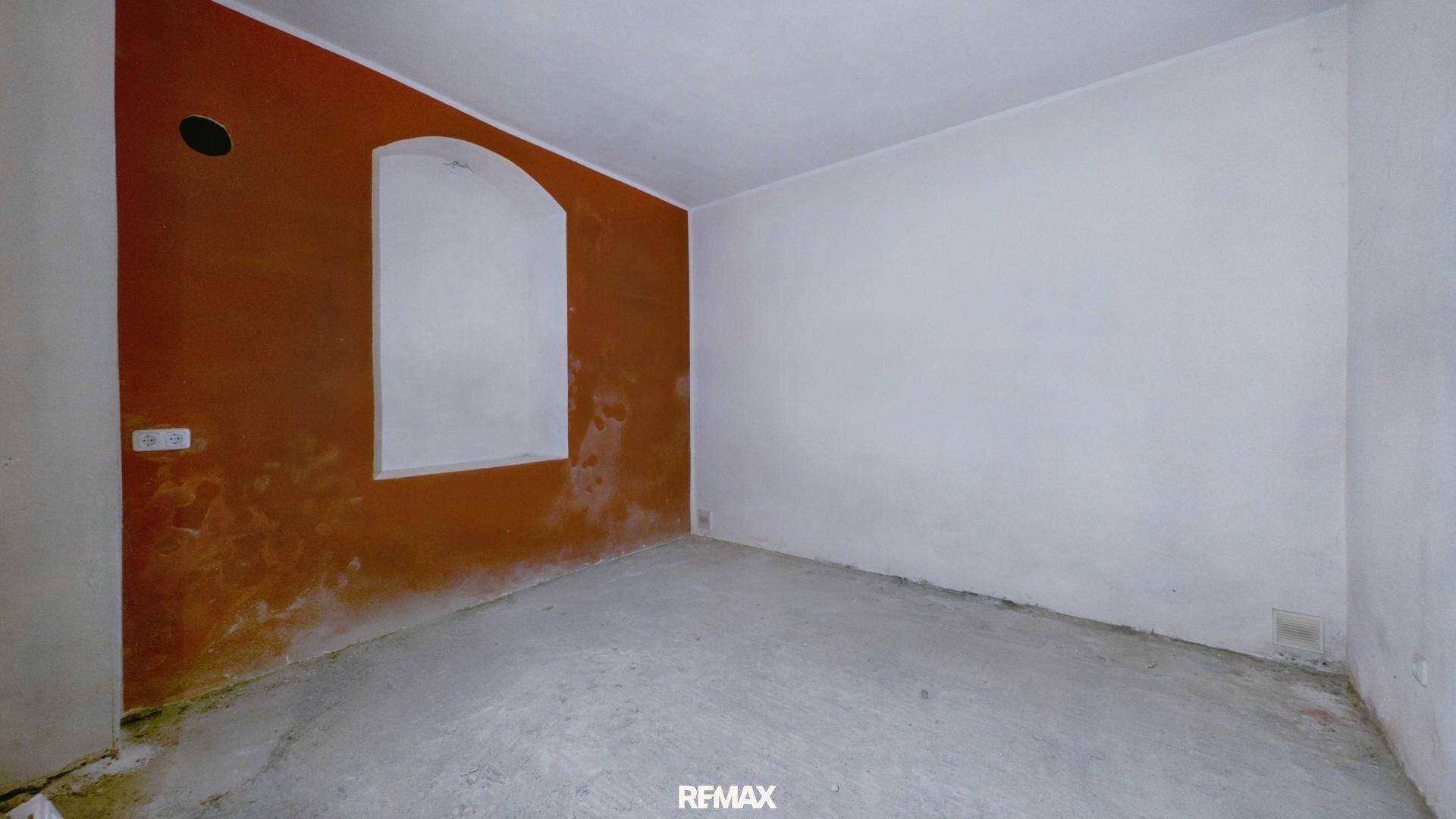 Renovierungsbedürftiger Raum mit einer orangefarbenen Wand und einem zugemauerten Bogenfenster.