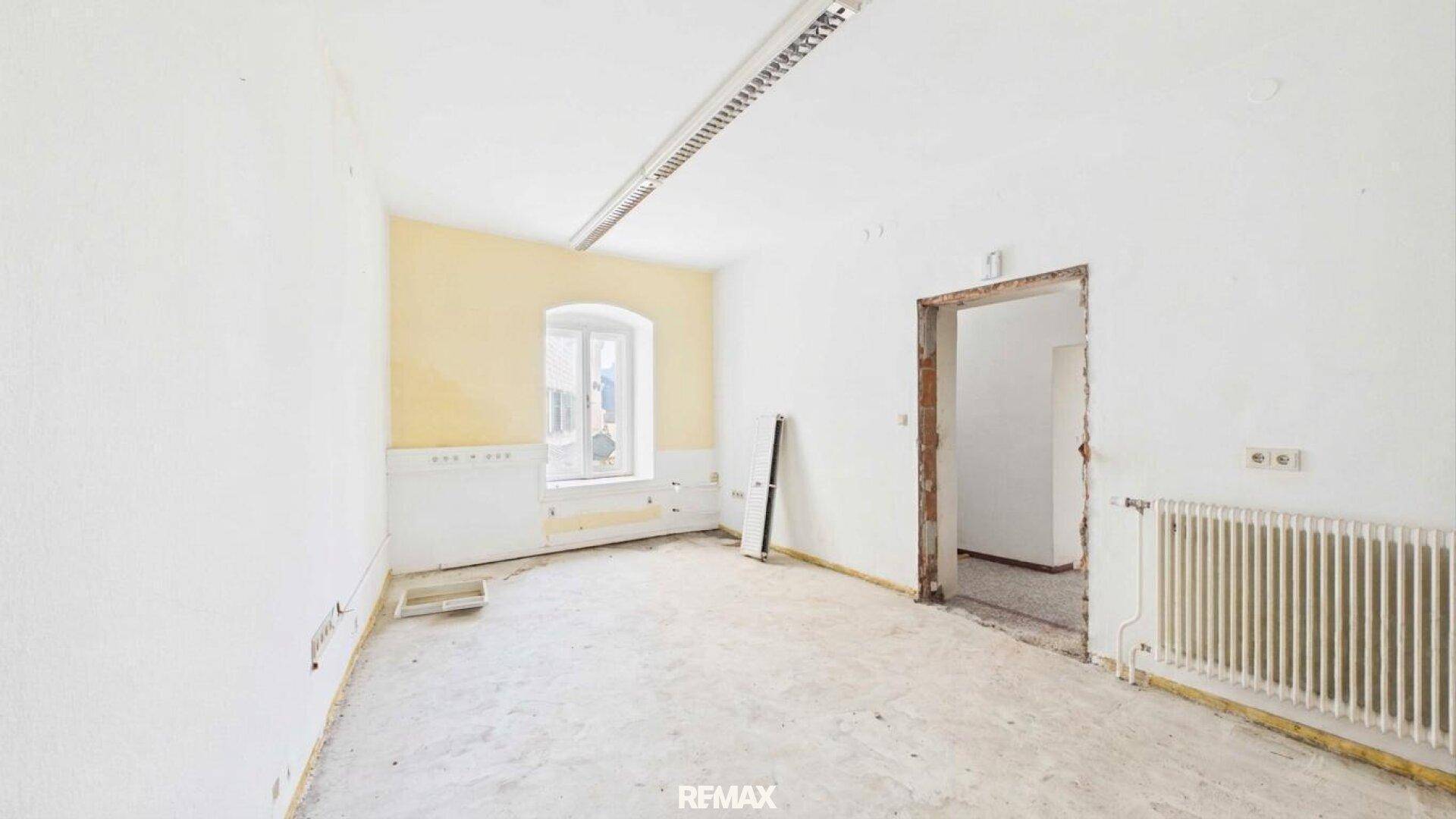 Renovierungsbedürftiger Raum mit gelber Wand, Fenster, Heizkörper und Deckenbeleuchtung.