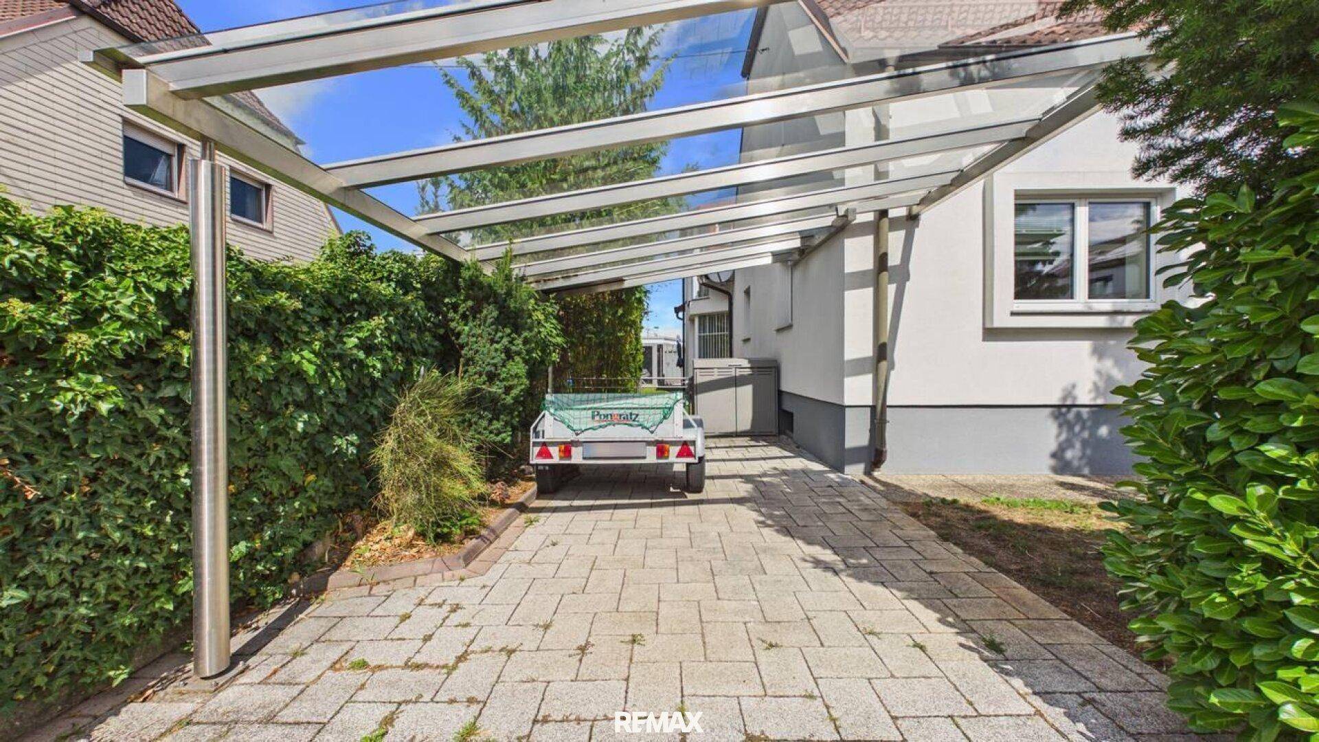 Moderner Carport mit Glasdach und gepflasterter Einfahrt, umgeben von Hecken.