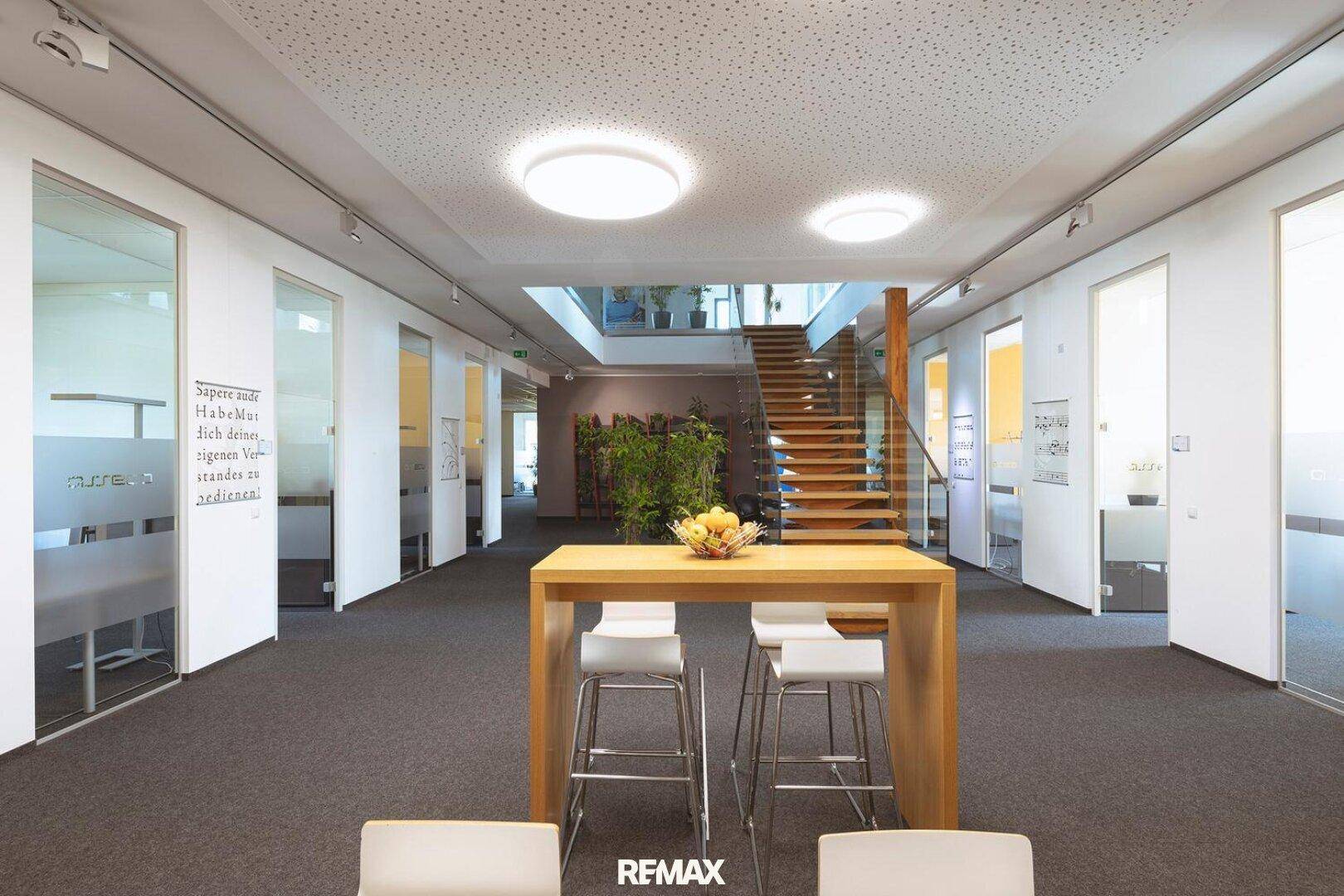 Offener Bürobereich mit Teppichboden, Glaswänden und einer Holztreppe, die in die obere Etage führt.